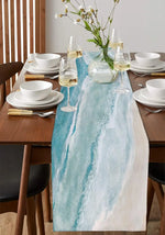 Cette table de salle à manger élégante est drapée d'une nappe bleue et blanche aux motifs aquatiques, complétée par des verres à vin, des assiettes blanches et un vase de fleurs sauvages, créant une atmosphère raffinée et apaisante.