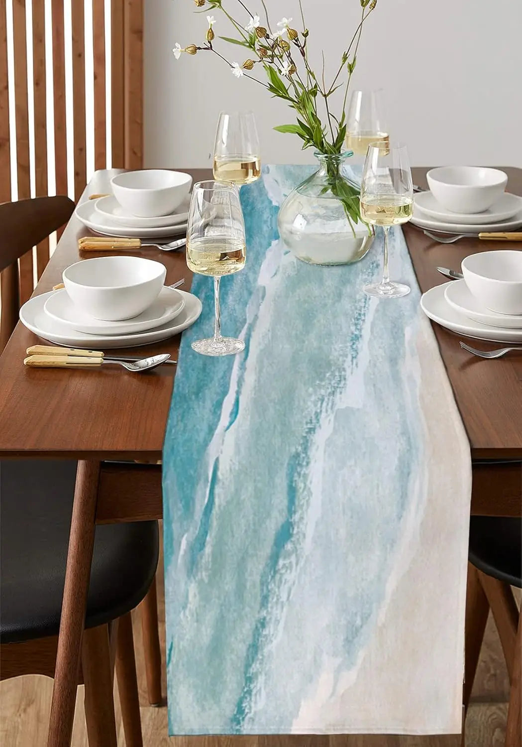 Cette table de salle à manger élégante est drapée d'une nappe bleue et blanche aux motifs aquatiques, complétée par des verres à vin, des assiettes blanches et un vase de fleurs sauvages, créant une atmosphère raffinée et apaisante.