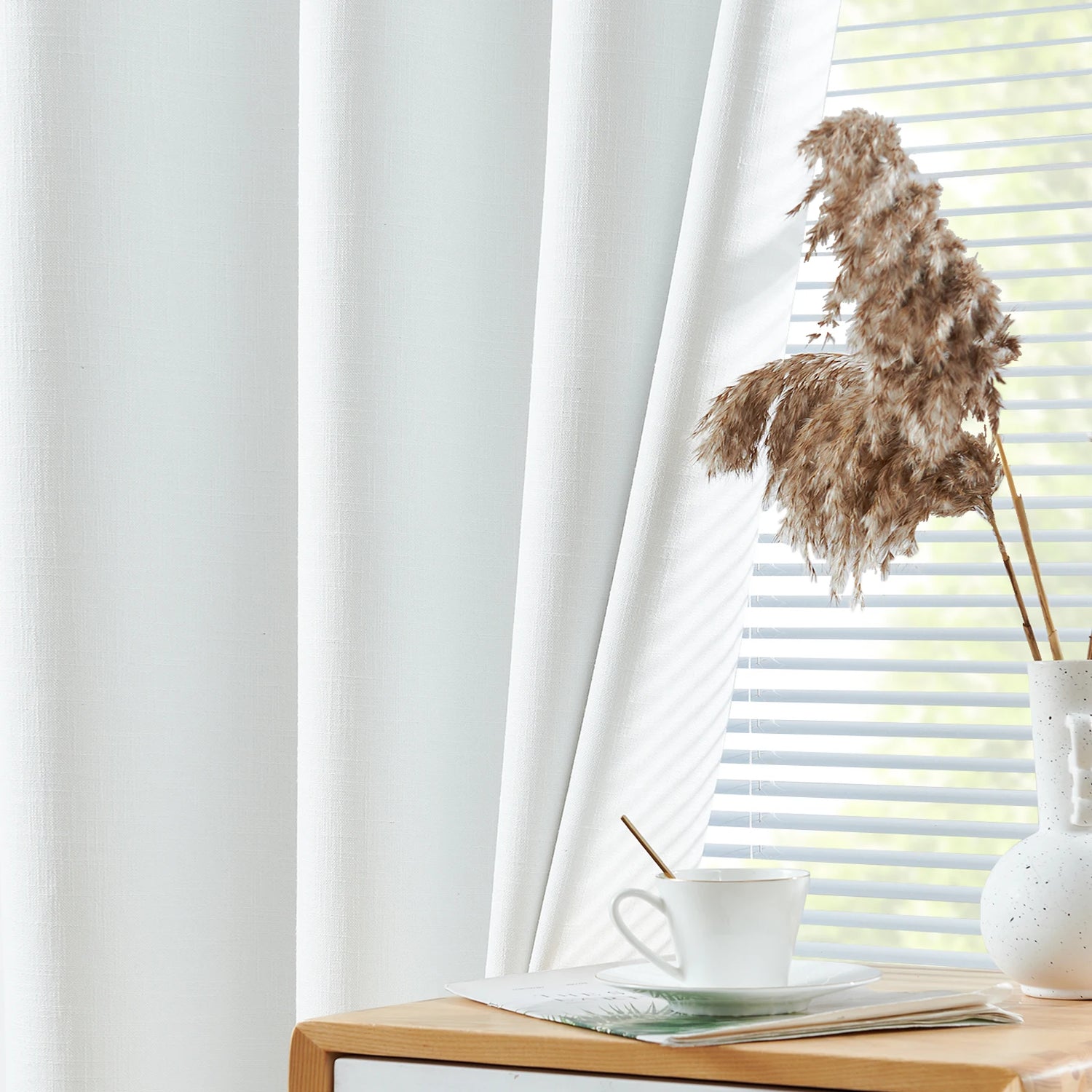 Rideau blanc élégant dans un salon moderne avec lumière naturelle. Vue de près sur une tasse de café posée sur une table en bois, créant une ambiance zen et apaisante pour la décoration intérieure. Fleurs séchées dans un vase minimaliste à côté des stores, accentuant l'esthétique chic et contemporaine de l'espace.