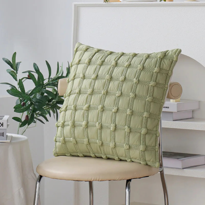 Coussin décoratif vert menthe à motifs géométriques carrés, placé sur une chaise blanche près d'une plante verte et de livres sur une étagère.