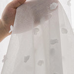 Détail d’un voilage blanc semi-transparent orné de petits motifs en relief, tenu à la main pour montrer la texture du tissu.