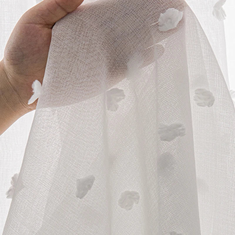 Détail d’un voilage blanc semi-transparent orné de petits motifs en relief, tenu à la main pour montrer la texture du tissu.