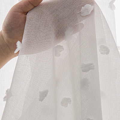 Détail d’un voilage blanc semi-transparent orné de petits motifs en relief, tenu à la main pour montrer la texture du tissu.