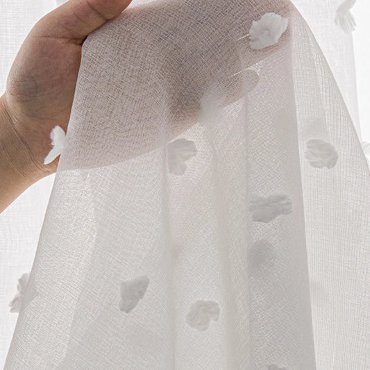 Détail d’un voilage blanc semi-transparent orné de petits motifs en relief, tenu à la main pour montrer la texture du tissu.