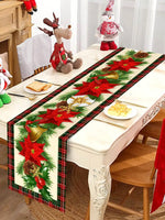 Chemin de table avec décoration de Noël : branchages de sapin, fleurs de poinsettia rouges, cloches dorées et nœuds sur un fond à carreaux rouges et verts.