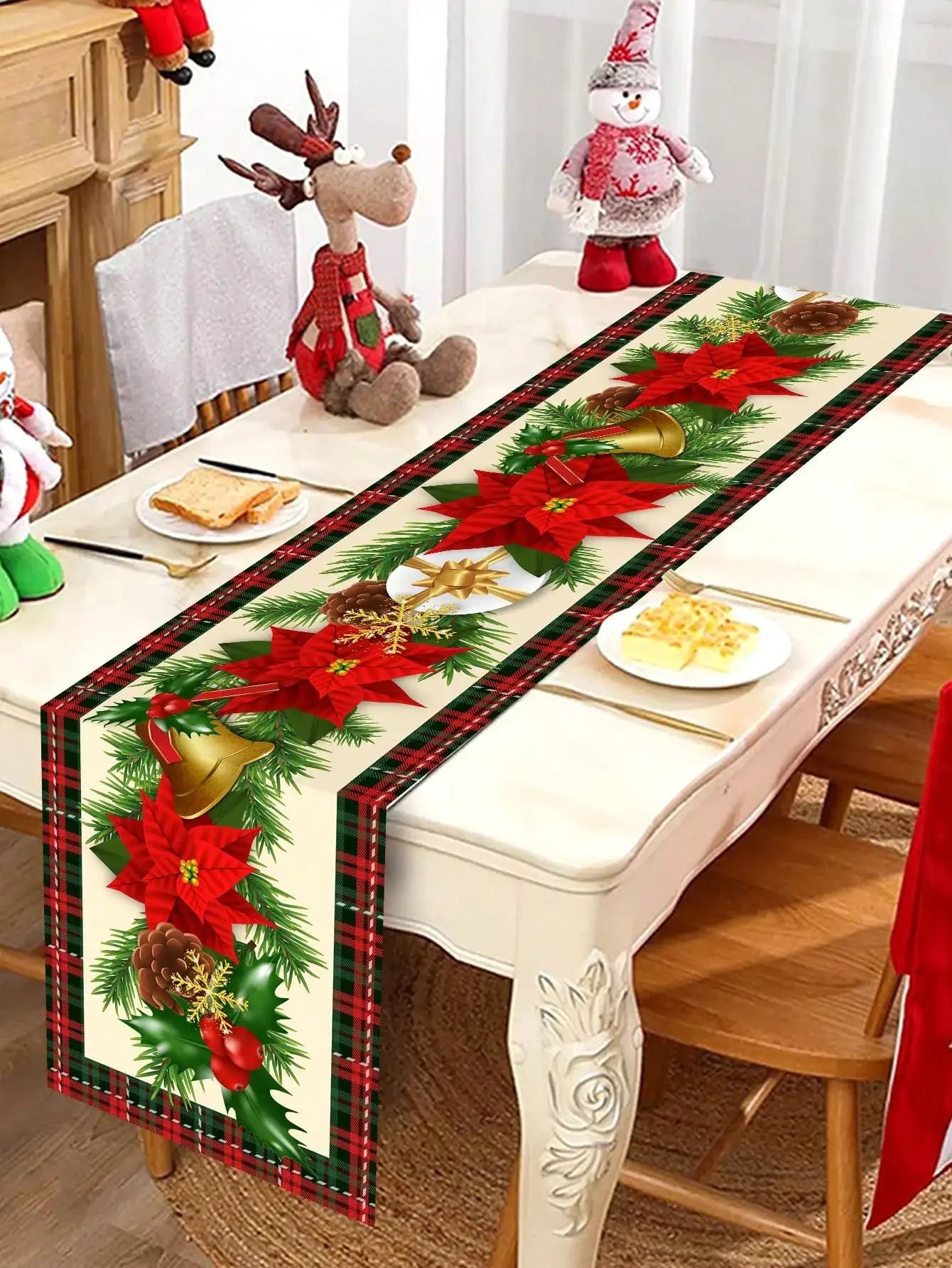 Chemin de table avec décoration de Noël : branchages de sapin, fleurs de poinsettia rouges, cloches dorées et nœuds sur un fond à carreaux rouges et verts.