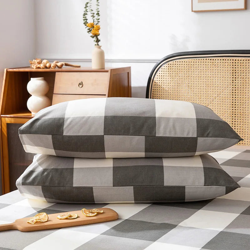 Deux oreillers gris et blancs carrés sur un lit avec une table de chevet en bois et un fauteuil en rotin dans une chambre confortable et lumineuse.