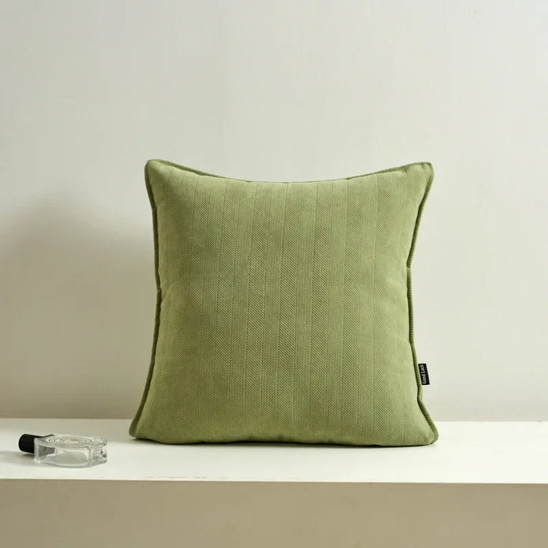 Un coussin décoratif vert olive, avec une texture douce et des lignes verticales, repose sur une surface blanche à côté d'un flacon en verre.