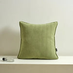 Un coussin décoratif vert olive, avec une texture douce et des lignes verticales, repose sur une surface blanche à côté d'un flacon en verre.
