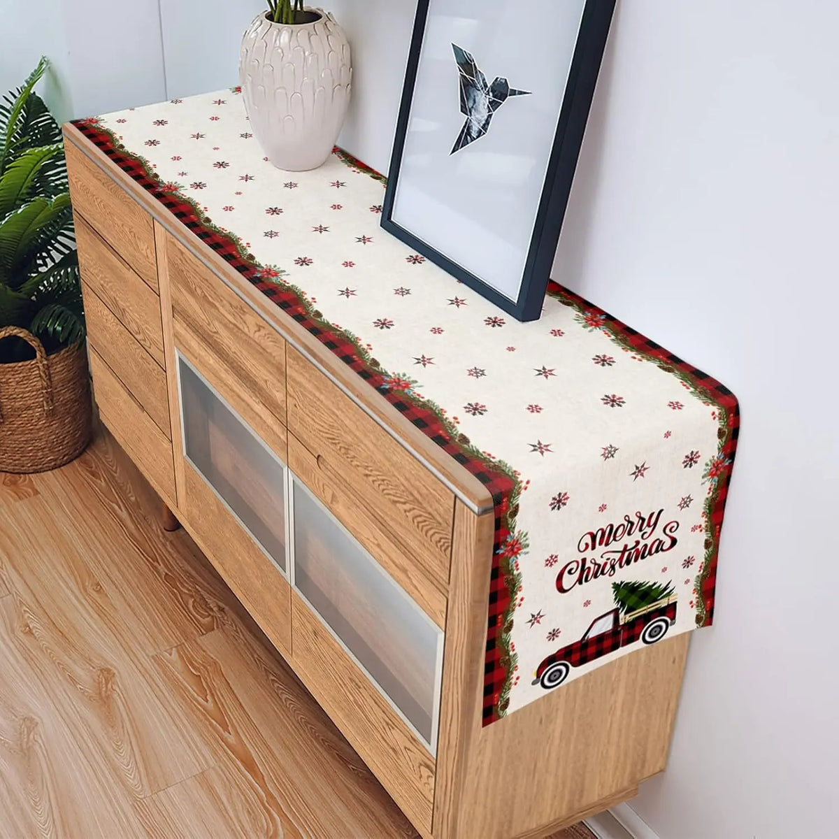 Sur un meuble en bois clair avec des tiroirs, un chemin de table aux motifs de flocons de neige et de voitures rouges vintage transmet une ambiance festive de Noël.