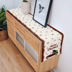 Sur un meuble en bois clair avec des tiroirs, un chemin de table aux motifs de flocons de neige et de voitures rouges vintage transmet une ambiance festive de Noël.