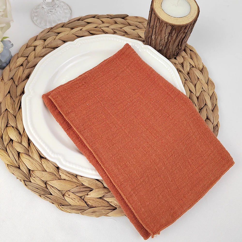 Une serviette de table en tissu de couleur rouille sur une assiette blanche posée sur un napperon en fibres tressées, entouré de bougies et d'un élément en bois.