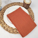 Une serviette de table en tissu de couleur rouille sur une assiette blanche posée sur un napperon en fibres tressées, entouré de bougies et d'un élément en bois.