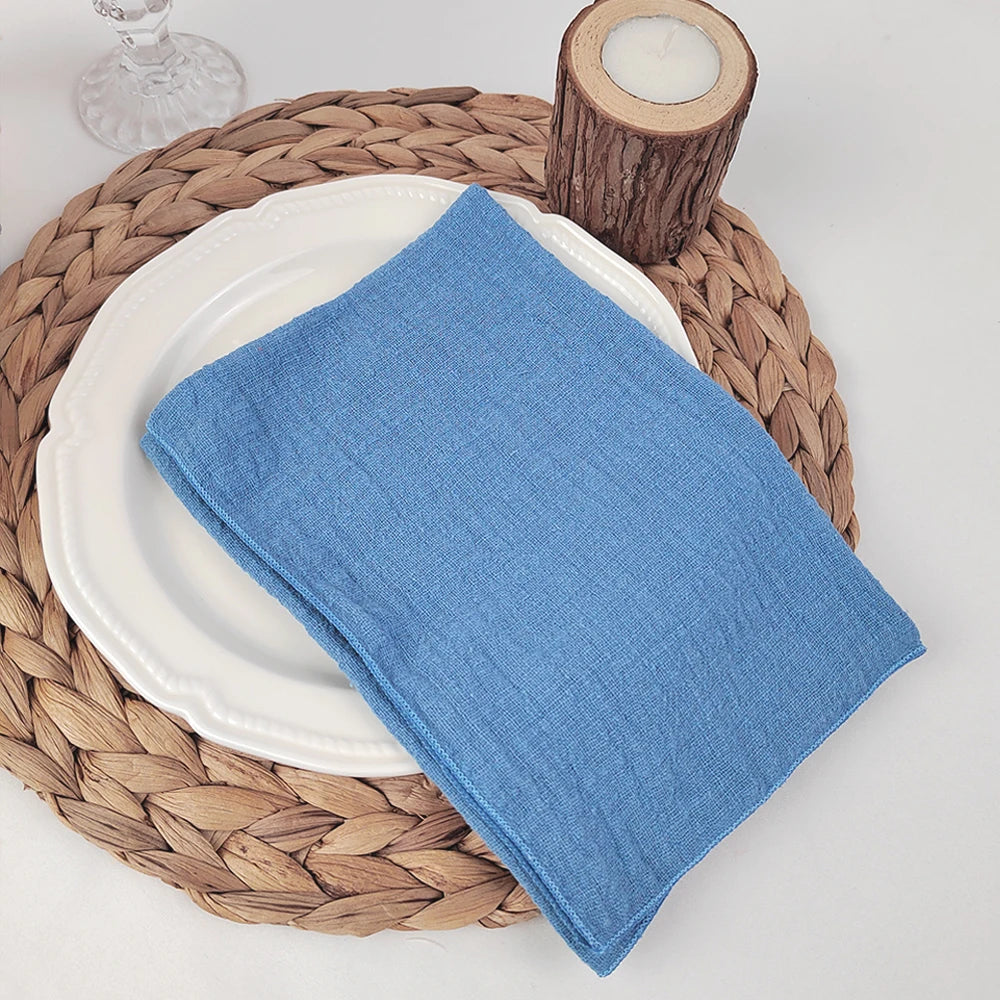 Serviette en lin bleu posée sur une assiette blanche sur un dessous de plat en osier tressé, accompagnée d'un photophore en bois.