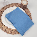 Serviette en lin bleu posée sur une assiette blanche sur un dessous de plat en osier tressé, accompagnée d'un photophore en bois.