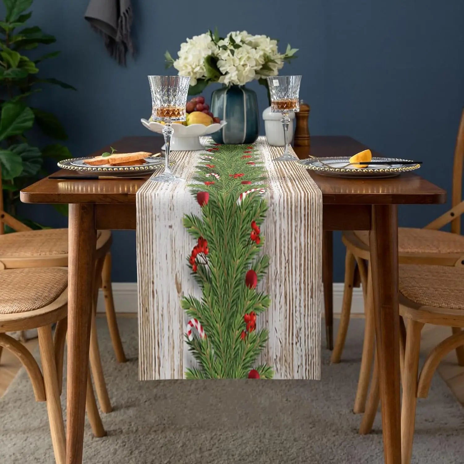 Une table rustique en bois avec un chemin de table en toile de jute décoré de brindilles de pins et de baies rouges, entourée de verres à vin et de fruits, créant une scène chaleureuse et festive.