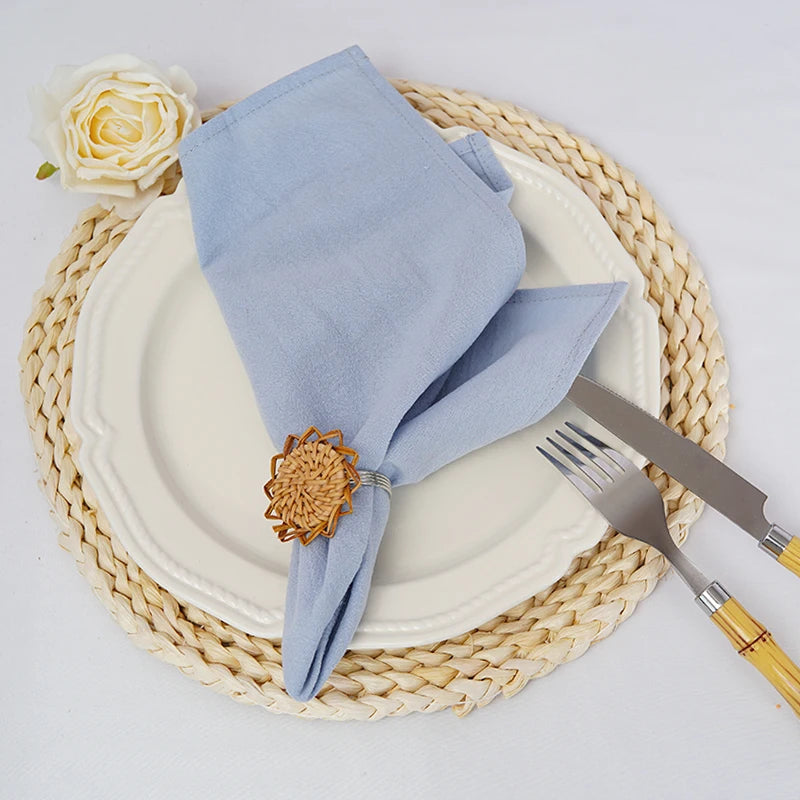 Une assiette blanche avec des couverts dorés est posée sur un tapis de paille beige, avec un linge de table bleu et une belle rose blanche.