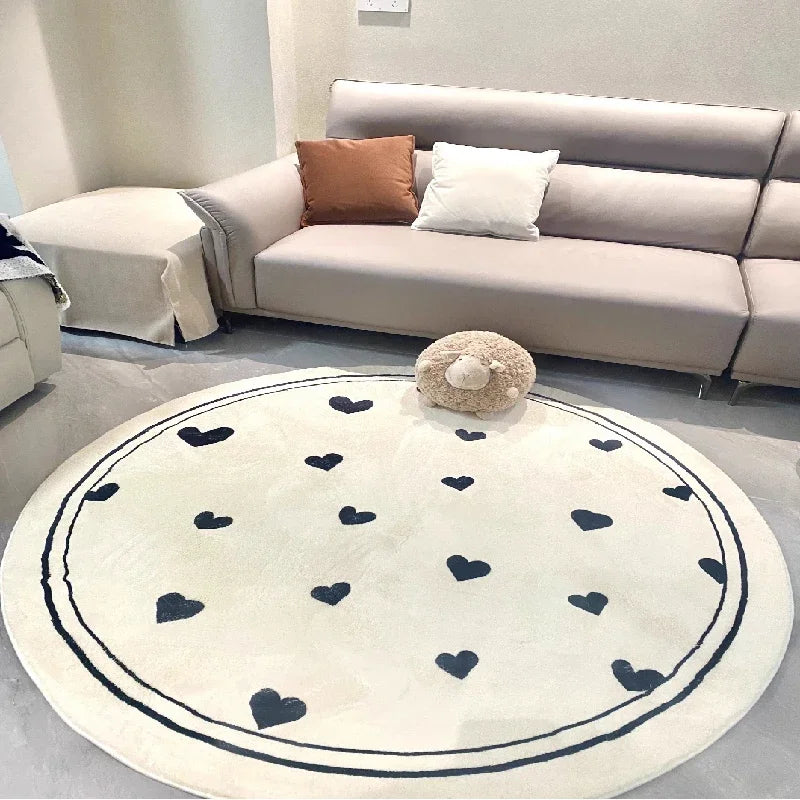 Tapis rond en peluche beige décoré de motifs de coeurs noirs, placé sur un sol dans un salon avec un canapé gris, des coussins et une peluche sur une table basse.