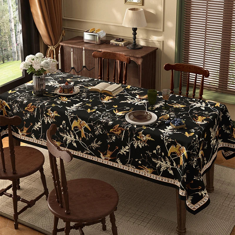 Cette image montre une salle à manger élégante avec une table recouverte d'une nappe noire ornée de motifs floraux et animaux, entourée de chaises en bois, et près d'un meuble vintage décoré de divers objets.
