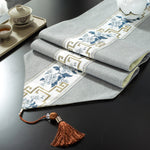 Gros plan de serviettes de table en lin gris ornées de motifs floraux et de bordures géométriques dorées, accompagnées d'un attache en raphia brun.
