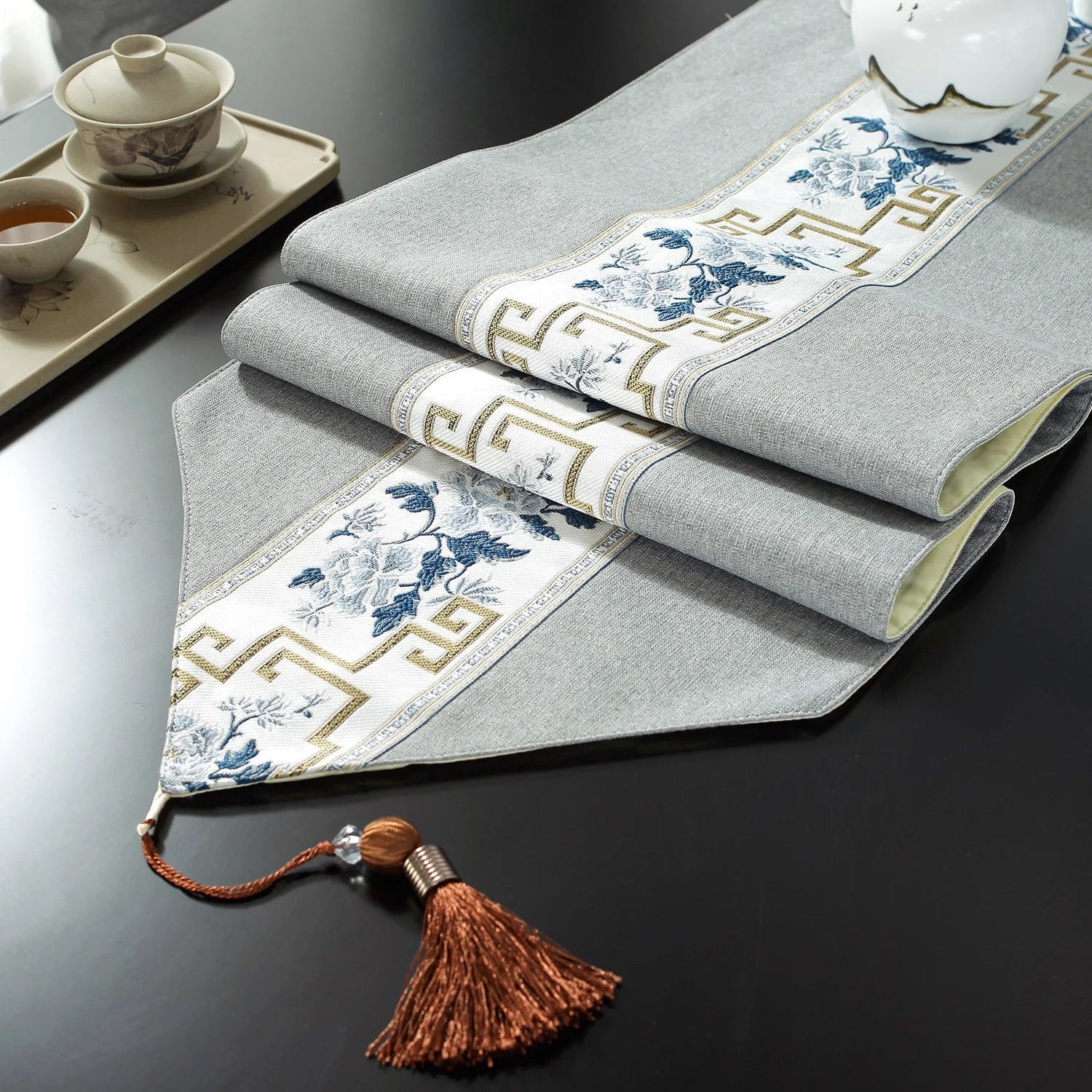 Gros plan de serviettes de table en lin gris ornées de motifs floraux et de bordures géométriques dorées, accompagnées d'un attache en raphia brun.