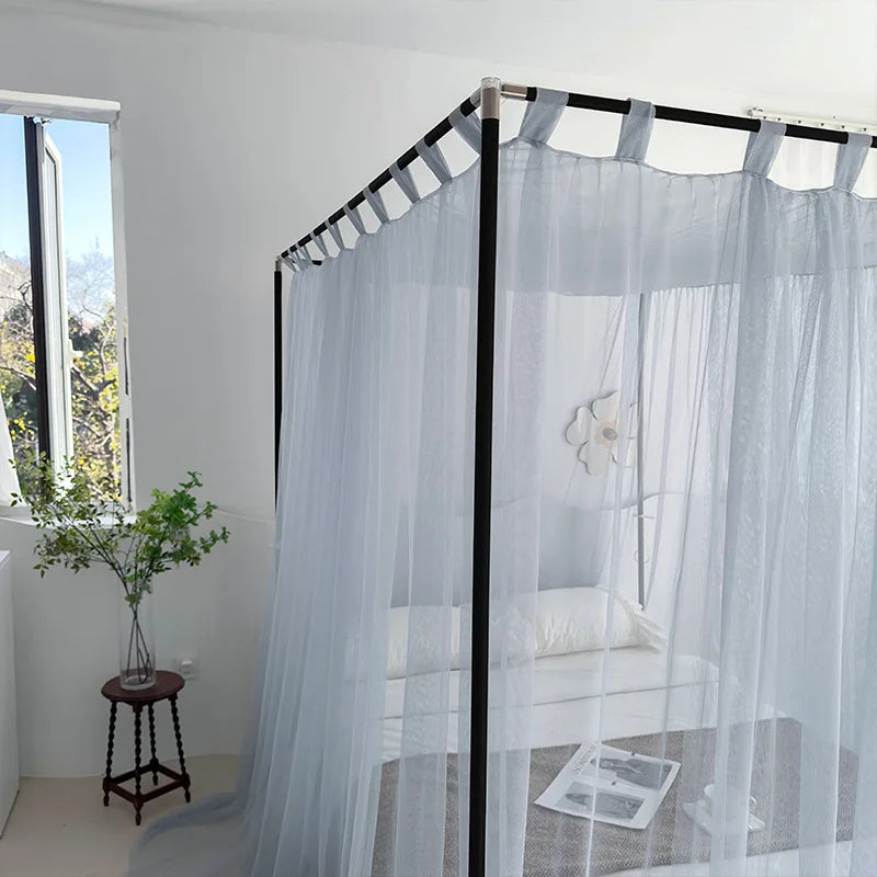 Un lit à baldaquin blanc dans une chambre lumineuse avec un tabouret en bois et une fenêtre laissant entrer la lumière naturelle.