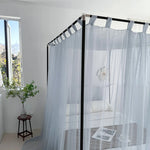 Un lit à baldaquin blanc dans une chambre lumineuse avec un tabouret en bois et une fenêtre laissant entrer la lumière naturelle.