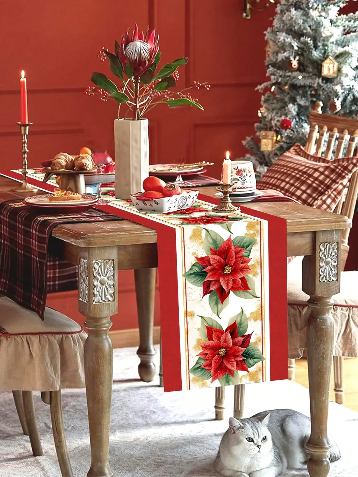 Une table de Noël richement décorée avec une nappe à carreaux, des bougies, une fleur de poinsettia dans un vase, et un chat gris endormi au premier plan, dans une atmosphère chaleureuse et festive.