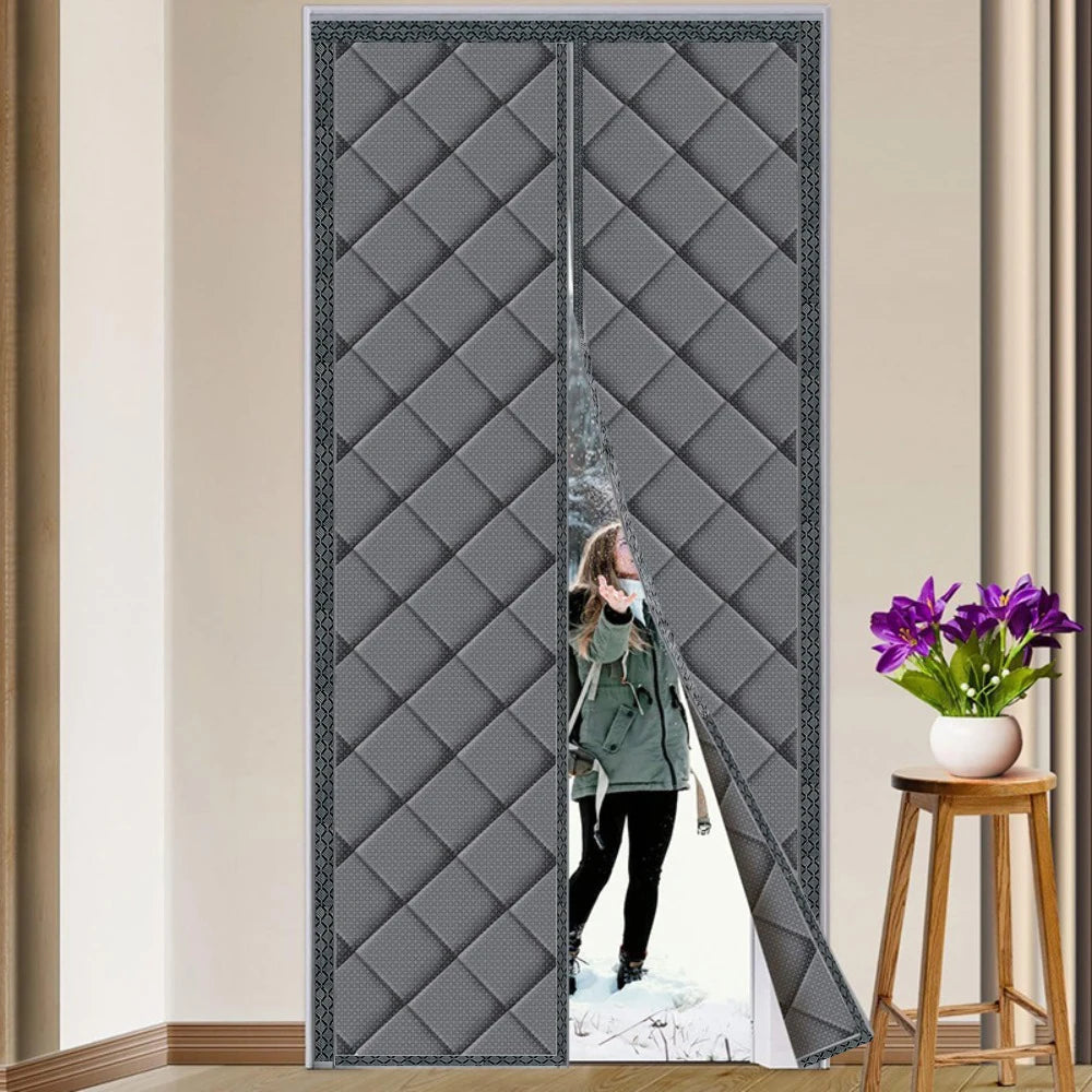 Femme en manteau d’hiver entrant dans une maison à travers un rideau isolant gris entrouvert, installé sur une porte intérieure, dans un décor sobre avec table d’appoint et fleurs.