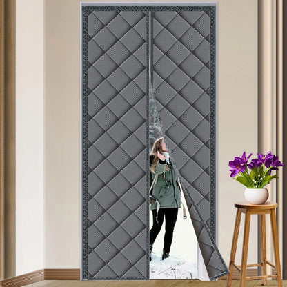 Femme en manteau d’hiver entrant dans une maison à travers un rideau isolant gris entrouvert, installé sur une porte intérieure, dans un décor sobre avec table d’appoint et fleurs.