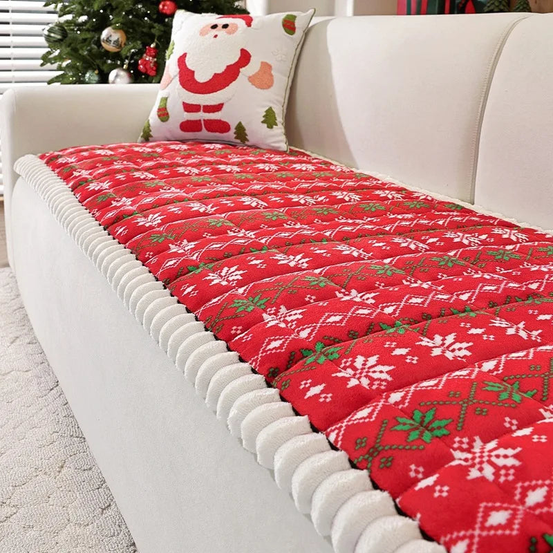 Un plaid en tricot rouge avec des motifs de Noël festifs, avec un coussin décoratif représentant le Père Noël, sur un canapé blanc à côté d'un sapin de Noël.