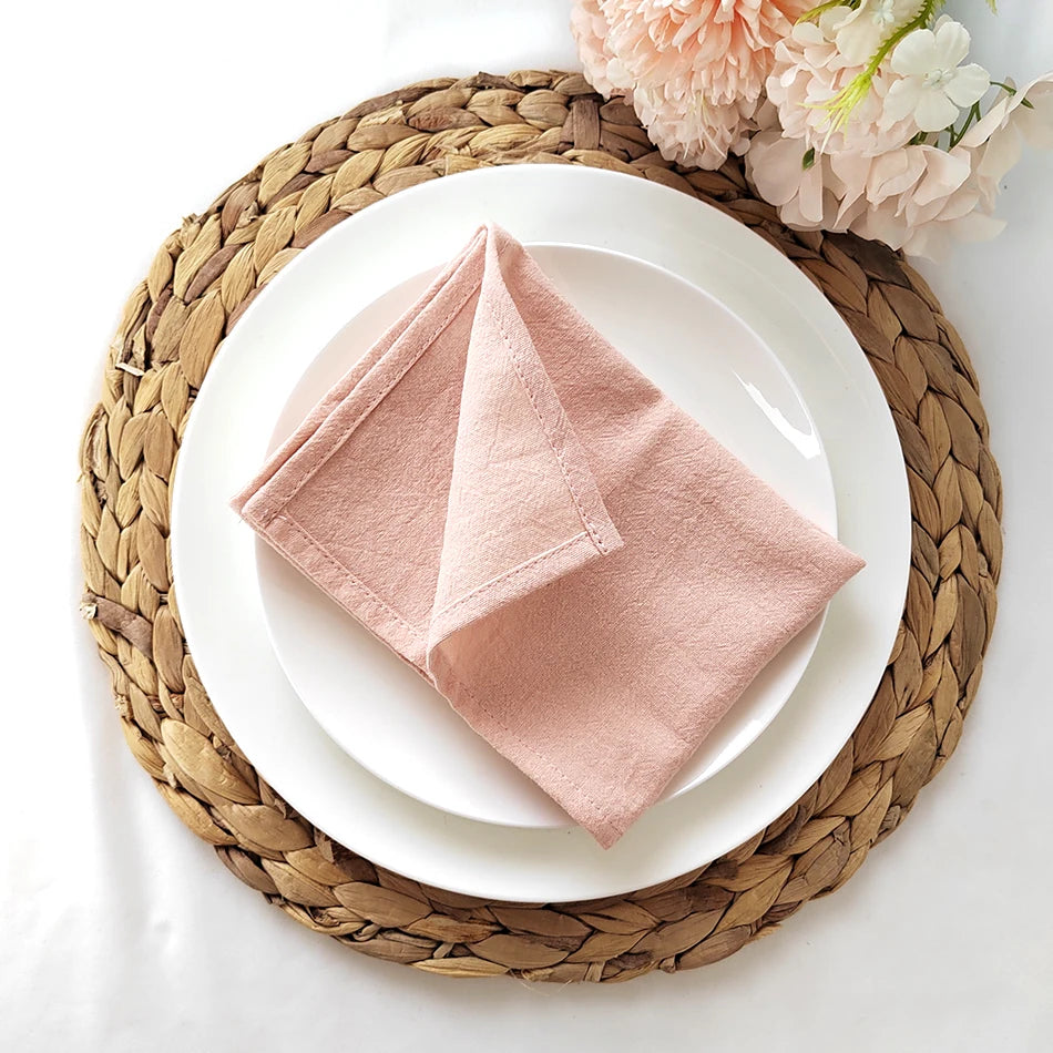 Une belle assiette blanche avec une serviette rose pâle pliée en forme de pyramide, posée sur un tapis de table rond tressé.