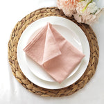 Une belle assiette blanche avec une serviette rose pâle pliée en forme de pyramide, posée sur un tapis de table rond tressé.