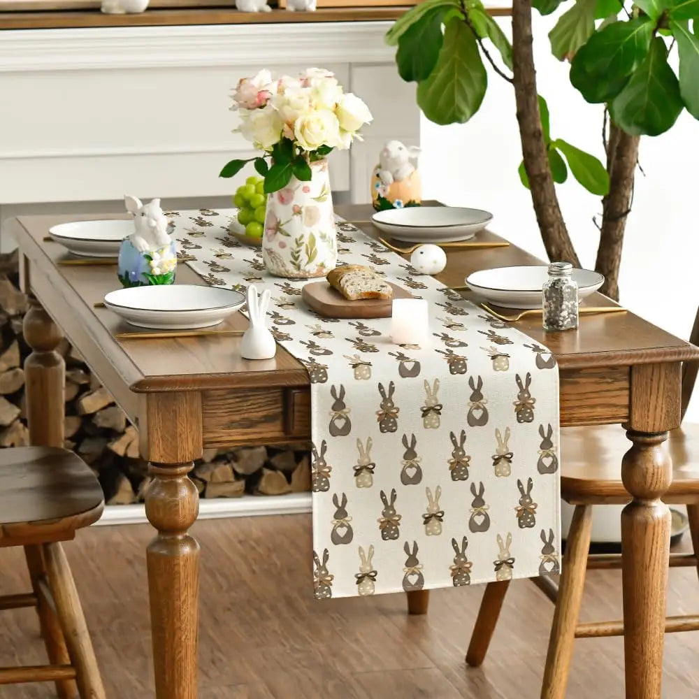Une table en bois rustique est joliment décorée avec un chemin de table motif lapins, des assiettes blanches et une jolie composition florale dans un vase orné, créant une atmosphère chaleureuse et printanière.
