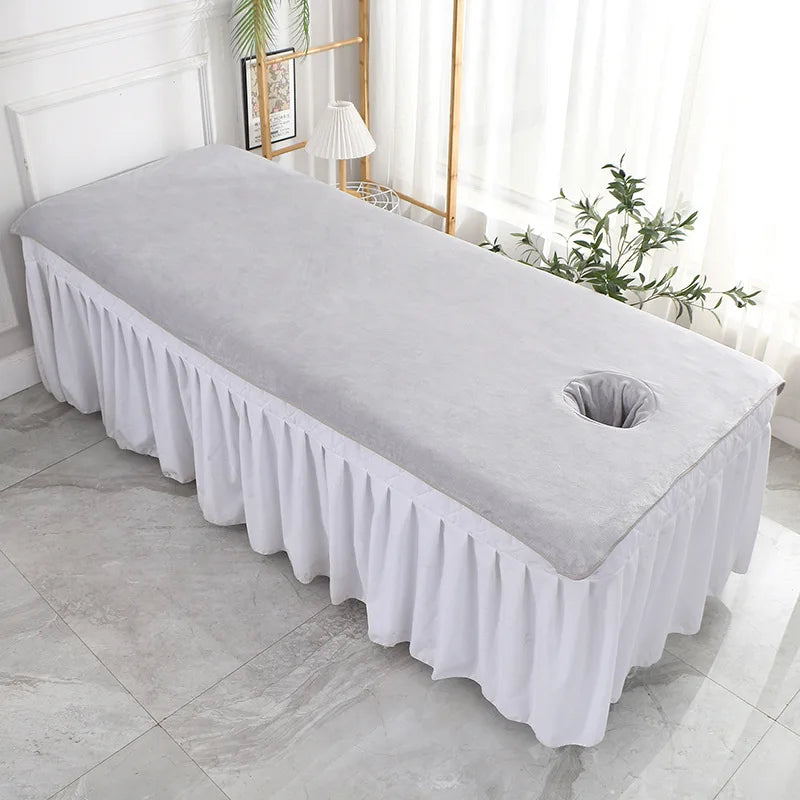 Cette image montre une table de massage recouverte d'un tissu blanc plissé, placée dans une pièce bien éclairée avec des rideaux blancs et une plante verte sur le côté.