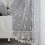 Voilage romantique blanc brodé suspendu sur une fenêtre, accompagné d’un meuble chic et d’un lampadaire plume, pour une ambiance raffinée et féminine.