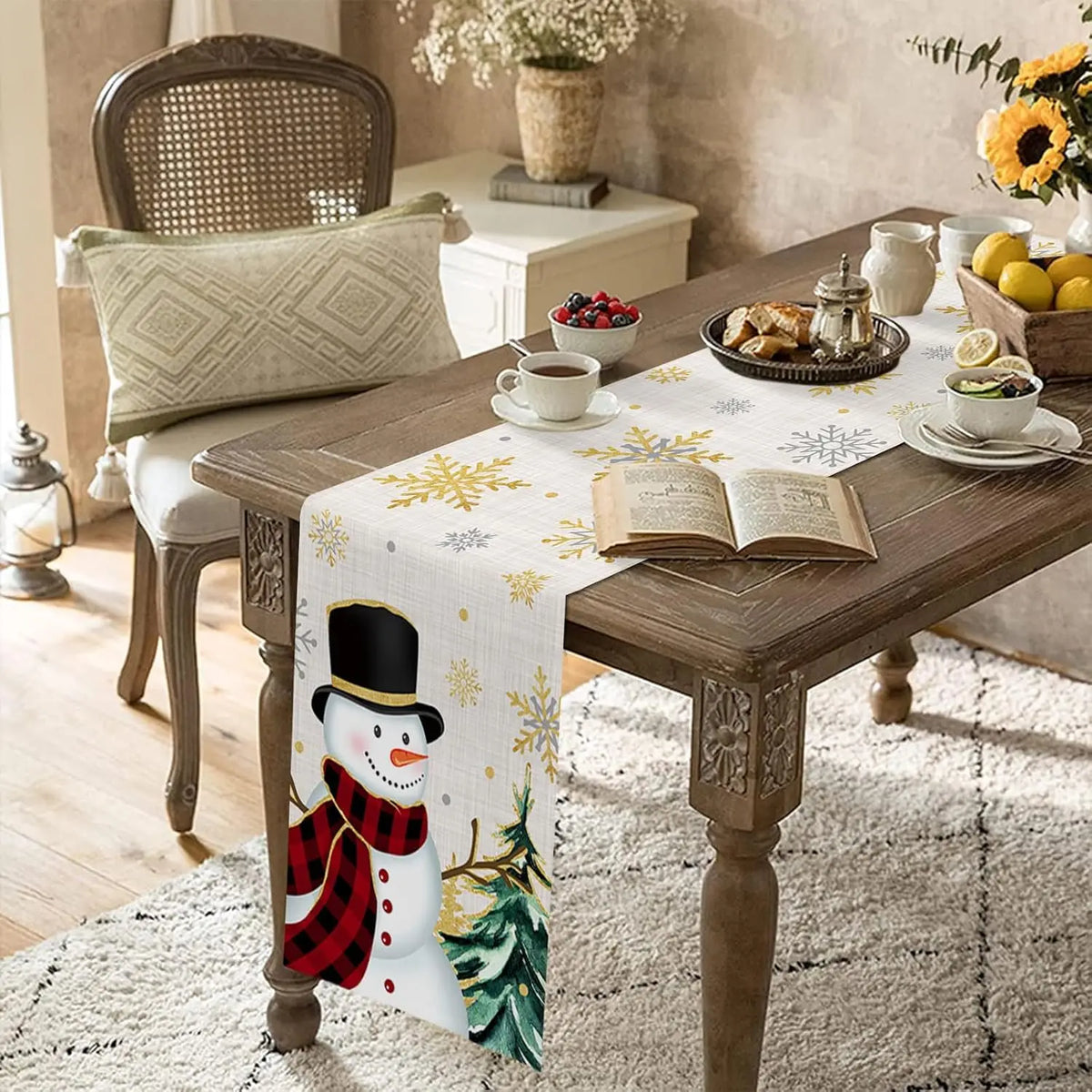 Une table de salon décorée pour Noël, avec une nappe imprimée de motifs hivernaux et un centre de table contenant des fleurs et des fruits. Un livre ouvert et une tasse de thé sont posés sur la table, créant une ambiance cozy et festive.