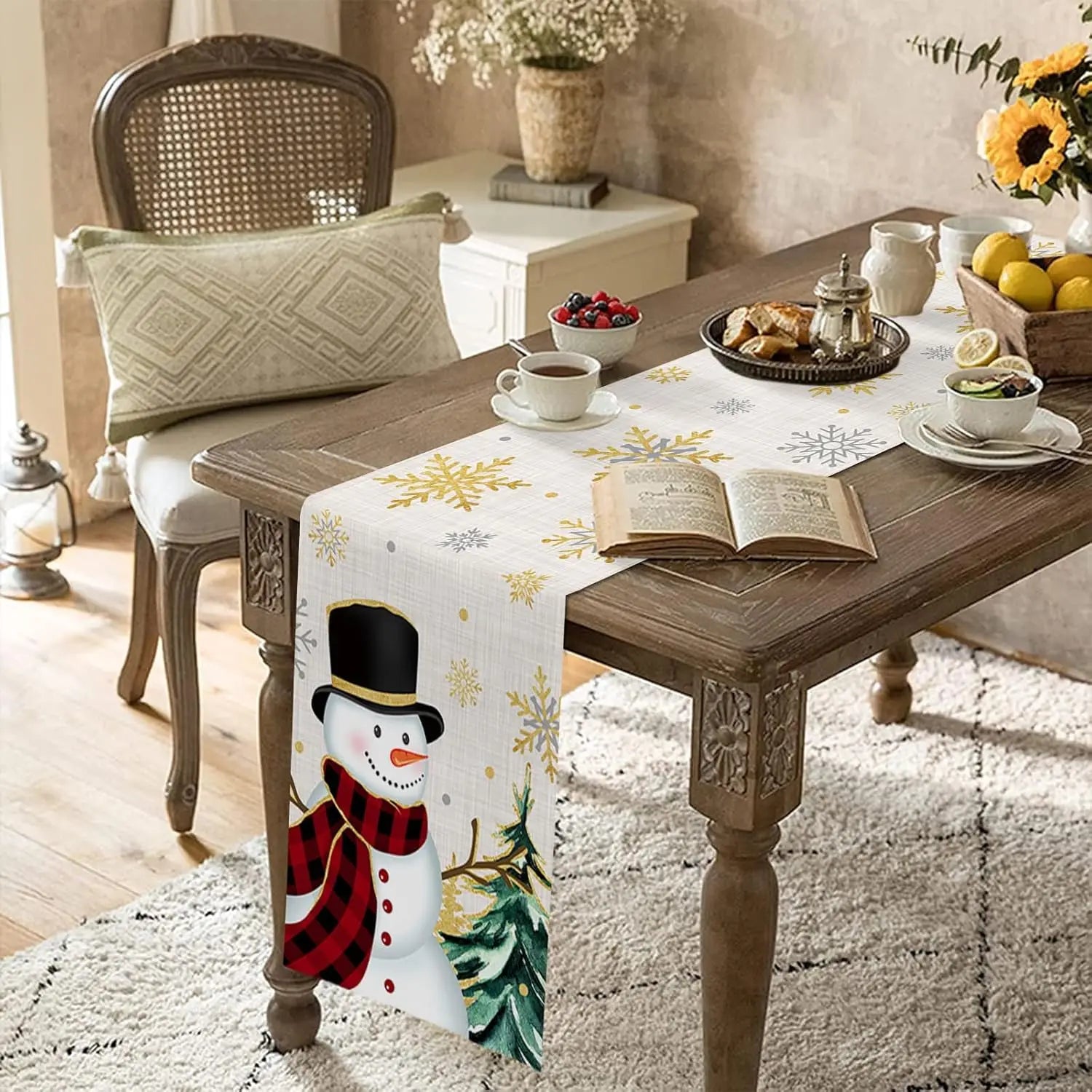 Une table de salon décorée pour Noël, avec une nappe imprimée de motifs hivernaux et un centre de table contenant des fleurs et des fruits. Un livre ouvert et une tasse de thé sont posés sur la table, créant une ambiance cozy et festive.