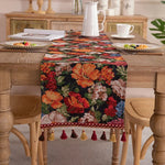 Cette image présente un chemin de table décoratif avec un motif floral coloré et vibrant, composé de fleurs oranges, rouges et roses sur un fond noir. Le chemin de table est orné de pompons et de franges qui ajoutent une touche de texture et de finition élégante à la décoration.