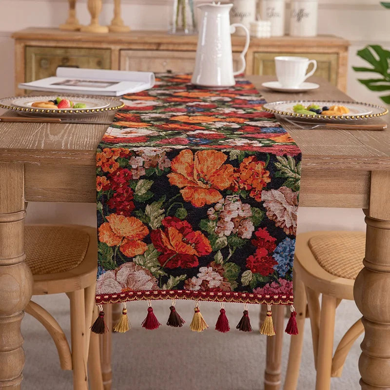 Cette image présente un chemin de table décoratif avec un motif floral coloré et vibrant, composé de fleurs oranges, rouges et roses sur un fond noir. Le chemin de table est orné de pompons et de franges qui ajoutent une touche de texture et de finition élégante à la décoration.