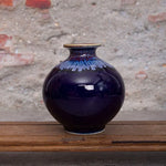 Un vase décoratif en céramique de couleur bleu foncé avec des nuances irisées, posé sur une surface en bois, offrant une élégance sophistiquée dans un style artisanal.