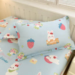 Image montrant un ensemble de literie pour enfant avec un motif de lapins et de fleurs sur un fond bleu clair. Des lapins blancs, des fraises et des fleurs sont représentés sur les oreillers et la couette, donnant à l'ensemble une apparence douce et ludique.