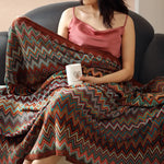 Femme vêtue d'un haut rose et d'un plaid coloré avec des motifs zigzags, tenant un objet blanc dans ses mains, assise sur un fauteuil sombre.