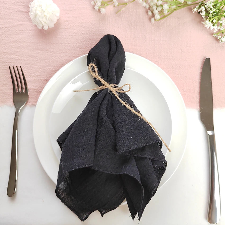 Un pliage de serviette noir enroulé avec de la ficelle, posé sur une assiette blanche avec des couverts, sur fond rose tendre avec des fleurs.