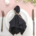 Un pliage de serviette noir enroulé avec de la ficelle, posé sur une assiette blanche avec des couverts, sur fond rose tendre avec des fleurs.