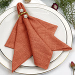 Une serviette de table pliée de couleur orange, ornée de perles blanches, posée sur une assiette blanche entourée de décorations végétales.