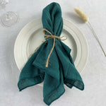 Serviette verte foncée pliée et attachée avec de la ficelle sur une assiette blanche, avec un verre et une brindille de plumes dans le fond.