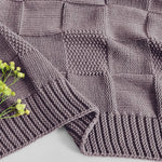 Un plaid tricoté à la main composé de panneaux carrés gris aux motifs entrelacés, rehaussé de quelques fleurs vertes aux extrémités.