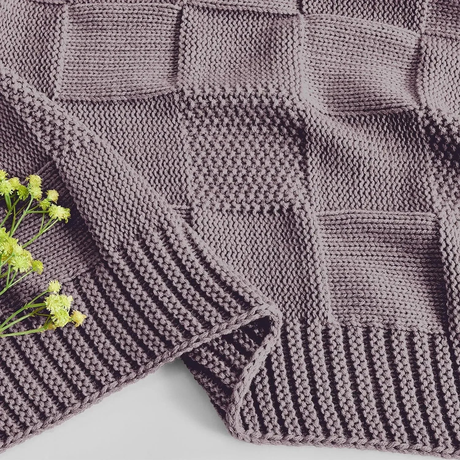 Un plaid tricoté à la main composé de panneaux carrés gris aux motifs entrelacés, rehaussé de quelques fleurs vertes aux extrémités.