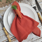 Serviette en tissu rouge enroulée et attachée avec un lien, décorée de brins de verdure verte, posée sur une assiette blanche avec des ustensiles en bois sur un fond de table en bois.